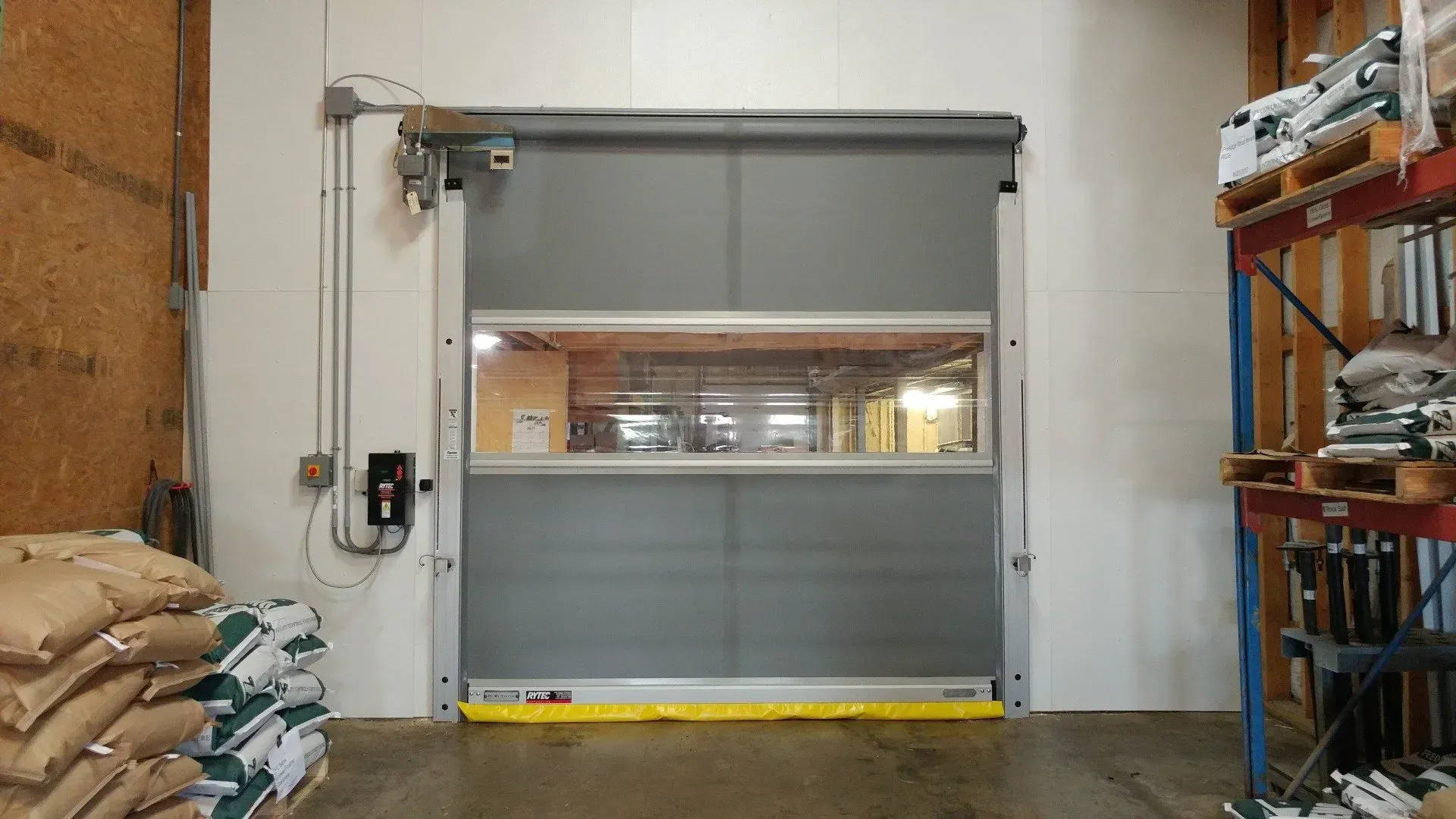 Grey high-speed industrial door, open to reveal a warehouse interior. Yellow safety strip at the bottom.