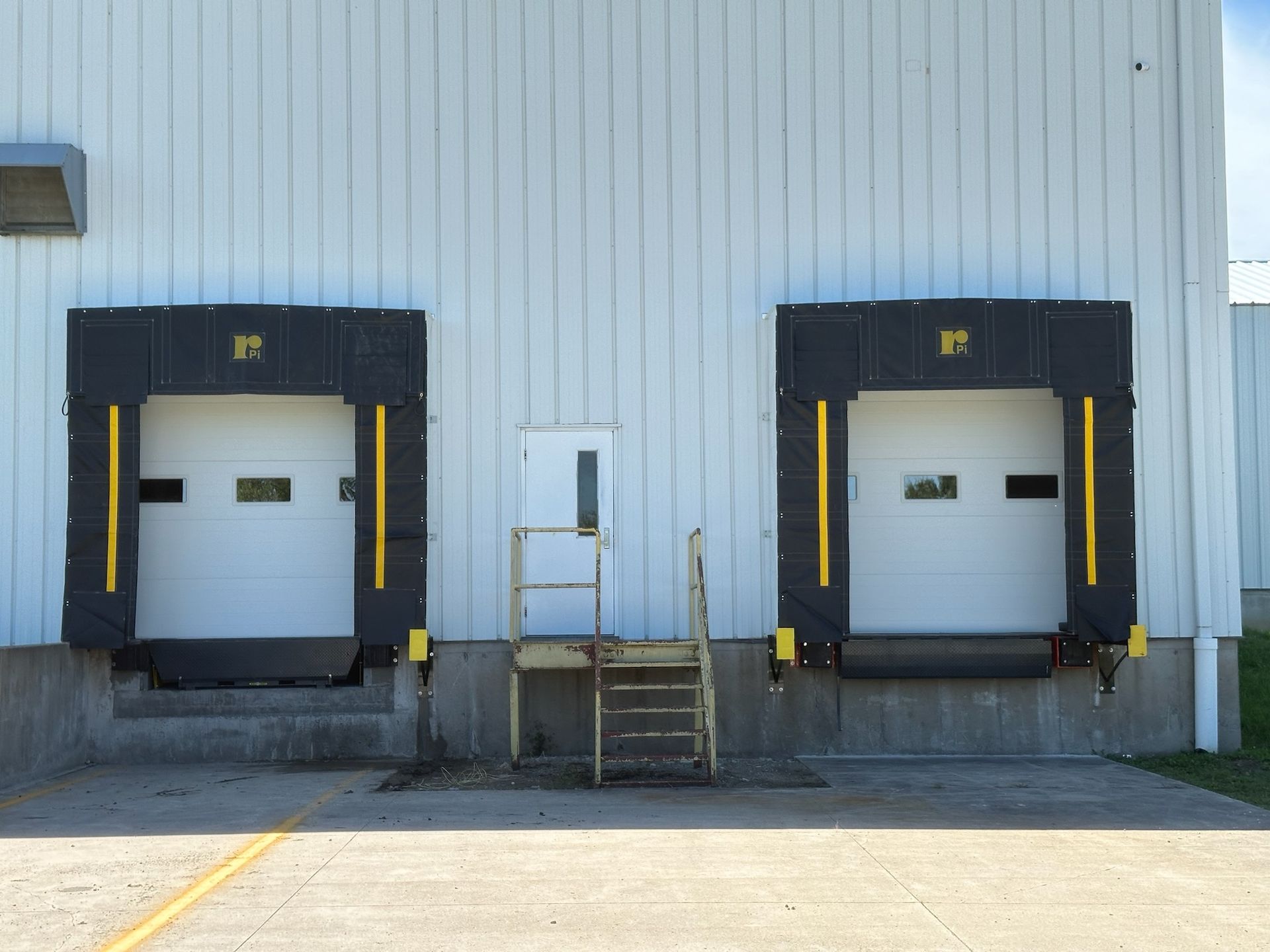 Two loading dock doors with black seals, white building. Small door in the middle with stairs.
