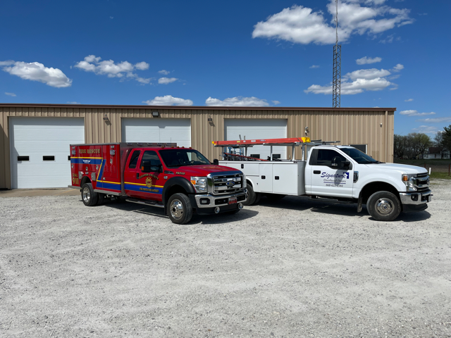 Two parked utility trucks, one red and one white, in front of a beige warehouse under a blue sky.