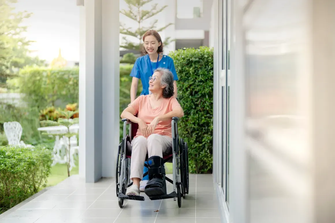 Caregiver pushing a woman in a wheelchair down a bright porch corridor with a garden view.