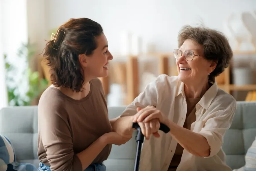 Two women smiling and talking on a couch, one holding a cane in a bright living room
