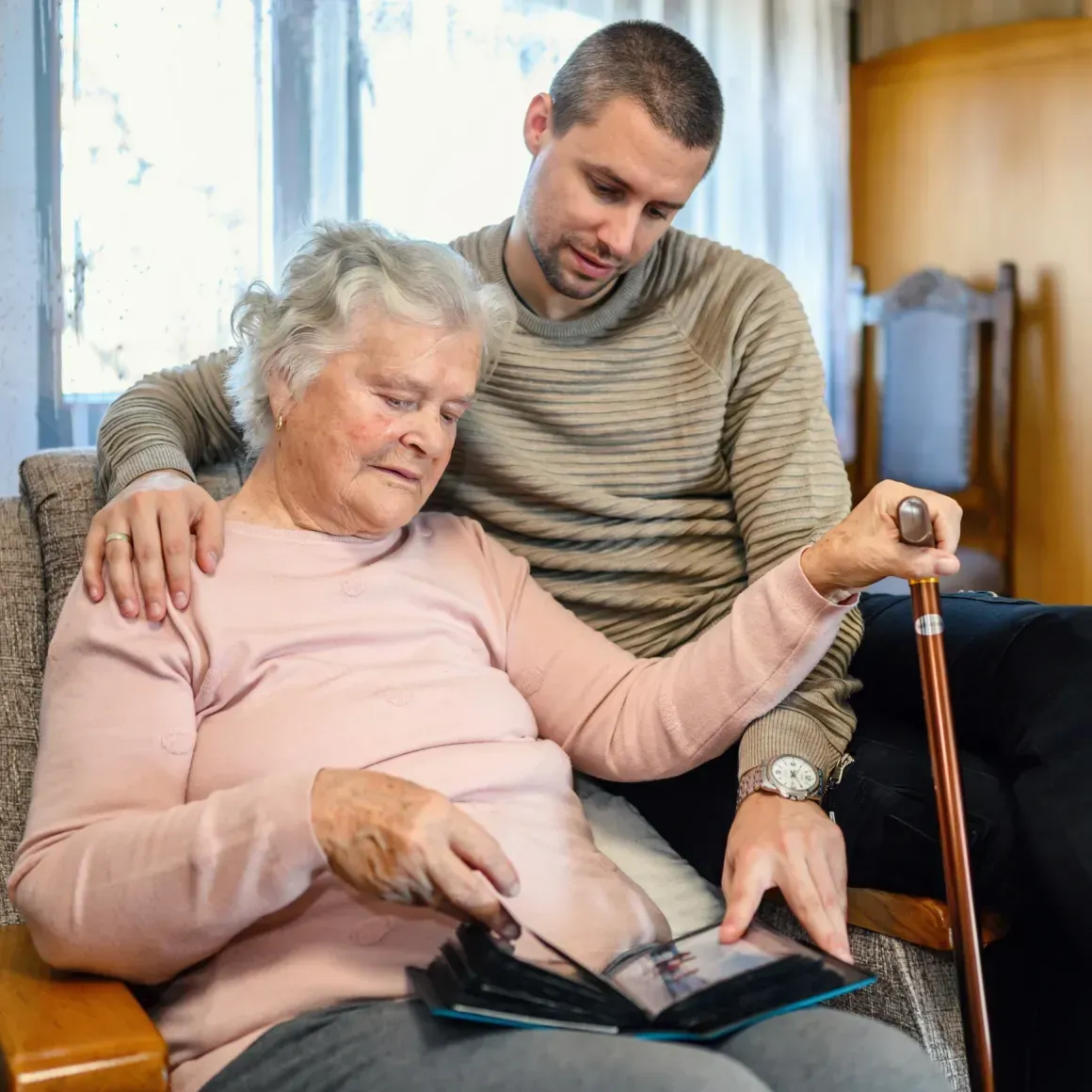 Man helping elderly woman read a book while seated on a couch in a cozy room