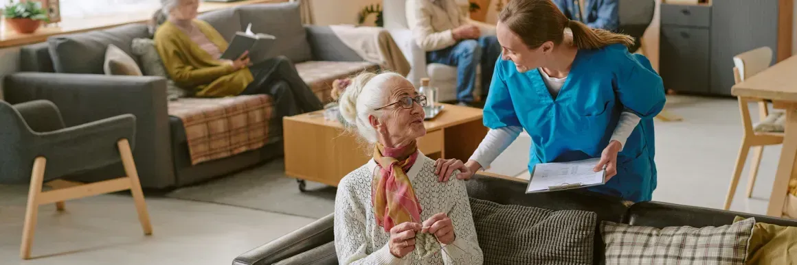 Caretaker helping an elderly person in a cozy living room while another person sits on a sofa.
