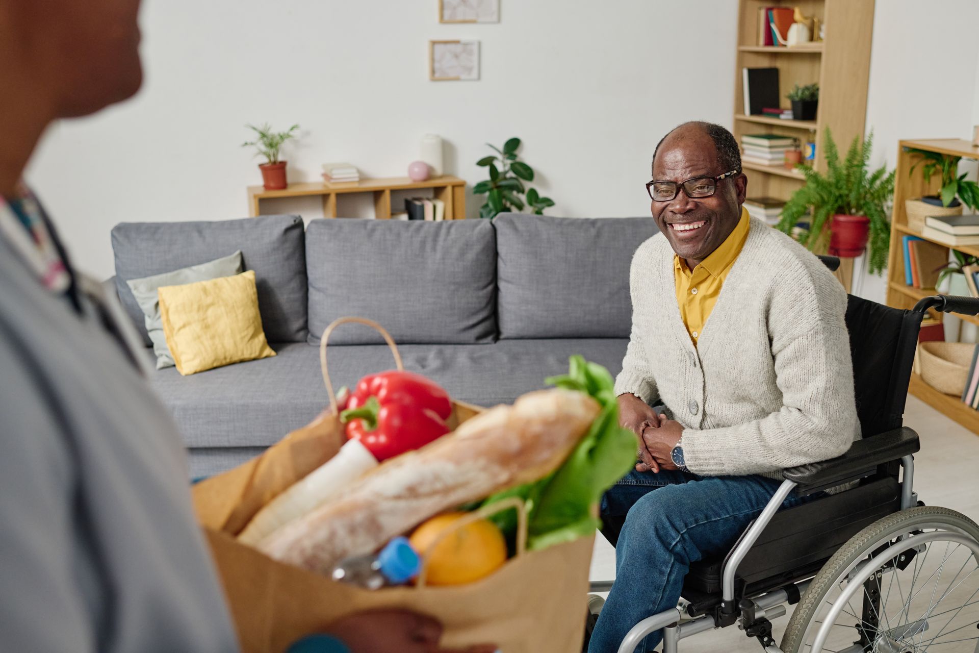 Person in a wheelchair smiles in a living room while receiving a grocery bag of fresh produce