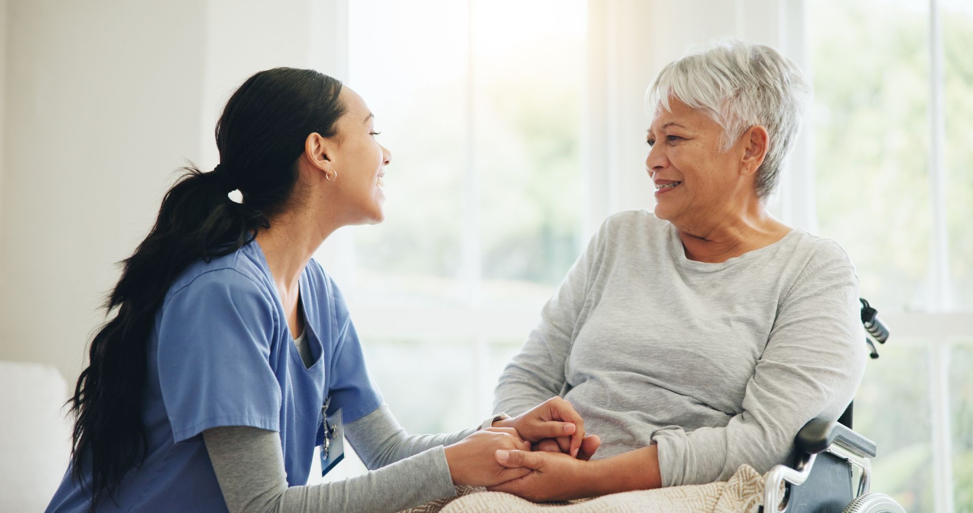 Caregiver in blue scrubs holding hands with a smiling patient in a bright room