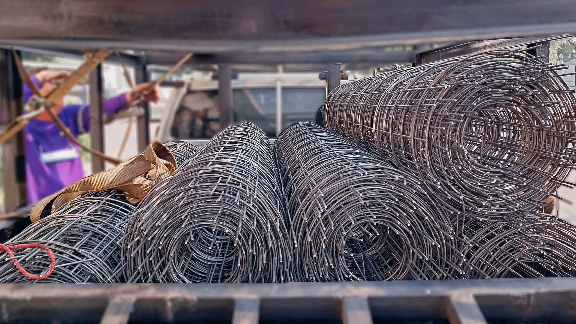 Coiled metal wire mesh rolls stacked in a tray, with purple straps and workshop background