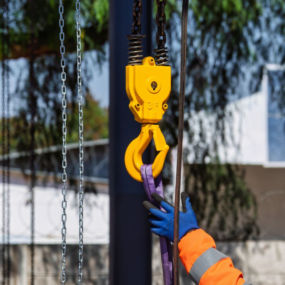 Yellow crane hook lifting a purple strap as a worker in blue gloves steadies it outdoors