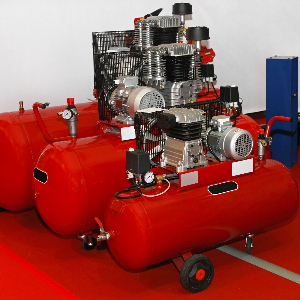 Two red industrial air compressors with silver motors and black cages on a workshop floor