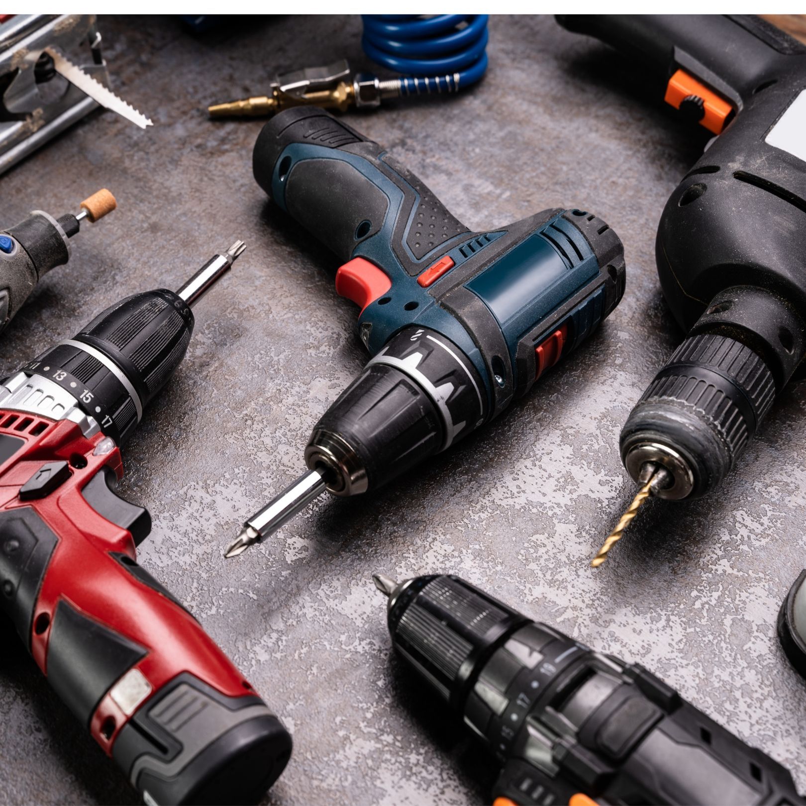 Assorted cordless drills and drill bits on a gray workbench, including red, black, and blue tools.