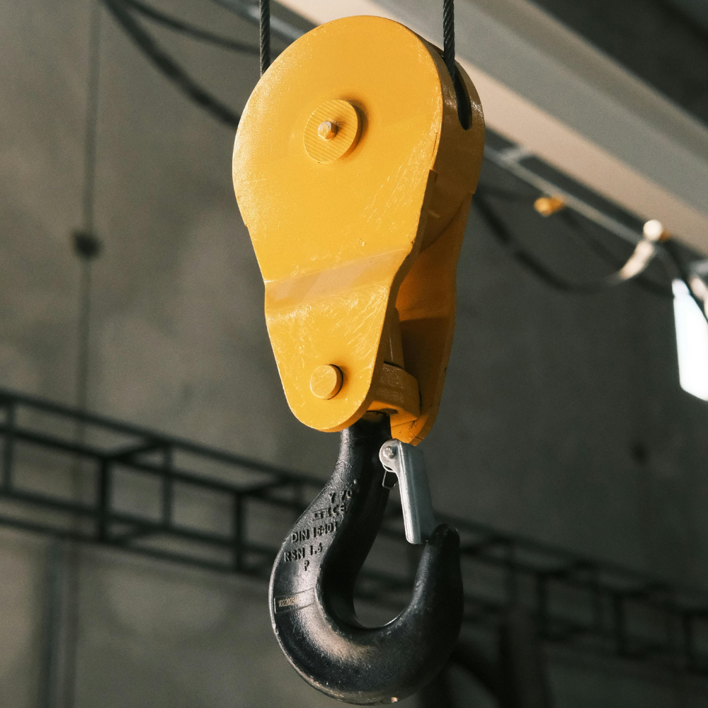 Yellow industrial crane hook hanging from a cable against a blurred indoor background