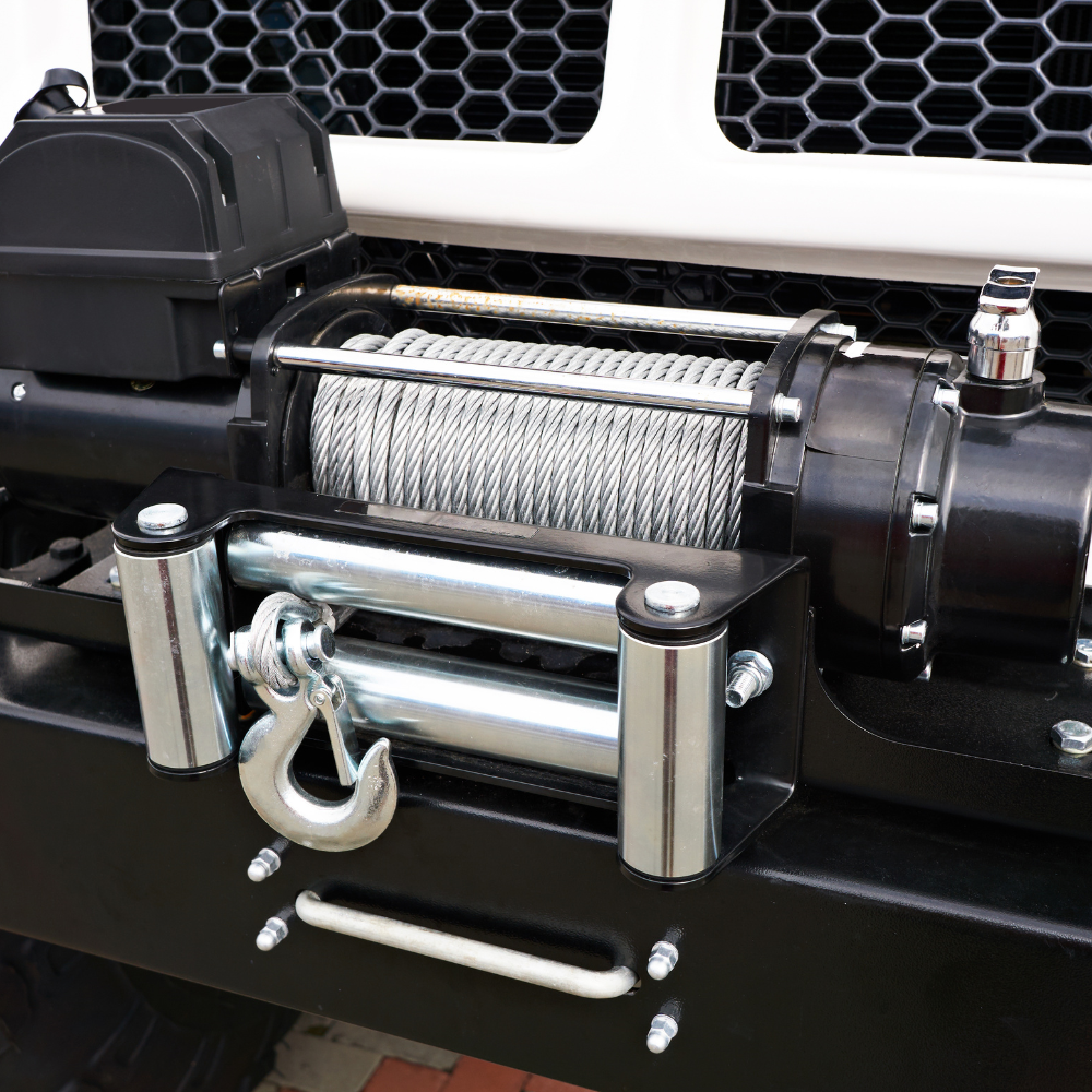 Close-up of a silver winch with steel cable, hook, and rollers mounted on a black vehicle bumper.