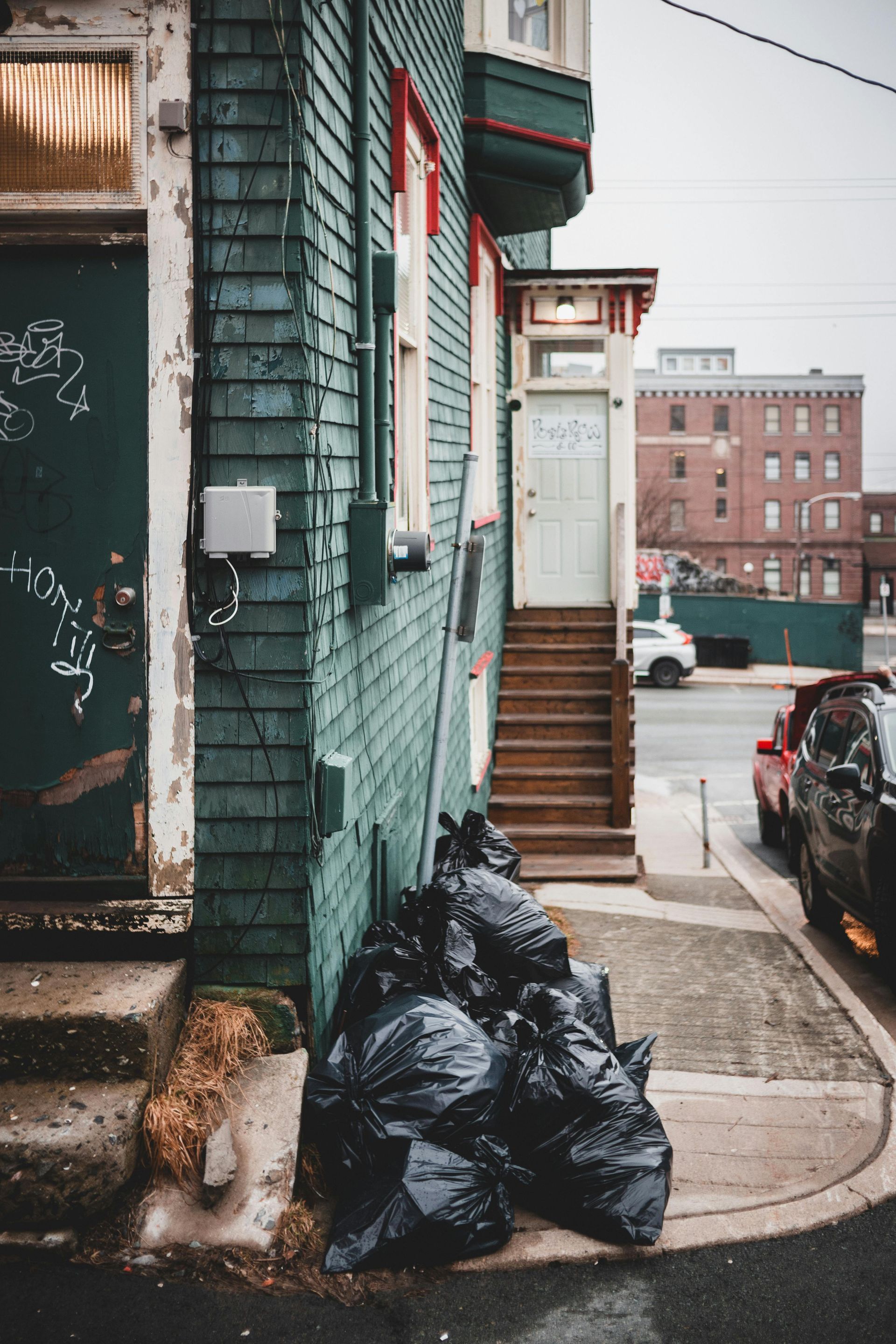 Trash bags piled on a city sidewalk beside a worn green building and stairway