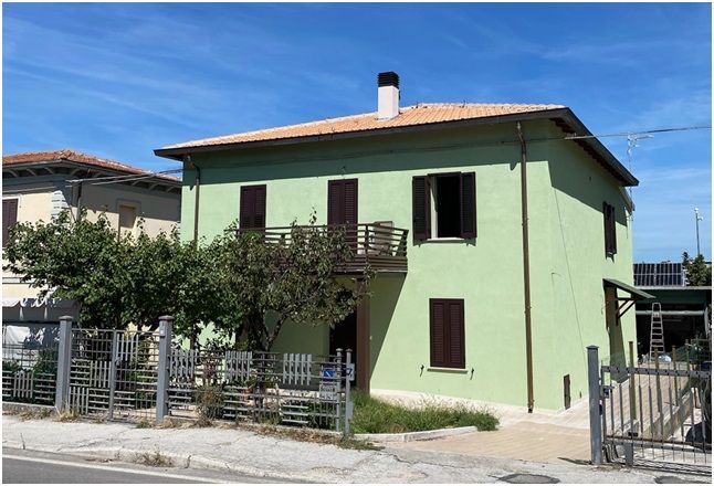 Casa verde a due piani con persiane marroni e balcone, situata in una strada soleggiata, sotto un cielo azzurro.