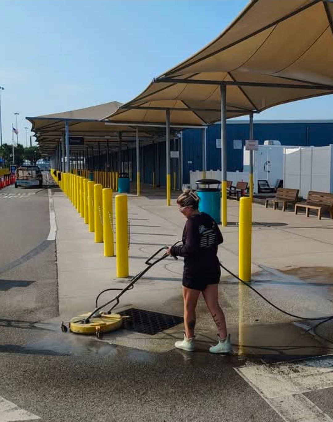 Worker power-washing a pavement beside yellow bollards and shaded walkway at a dock or terminal