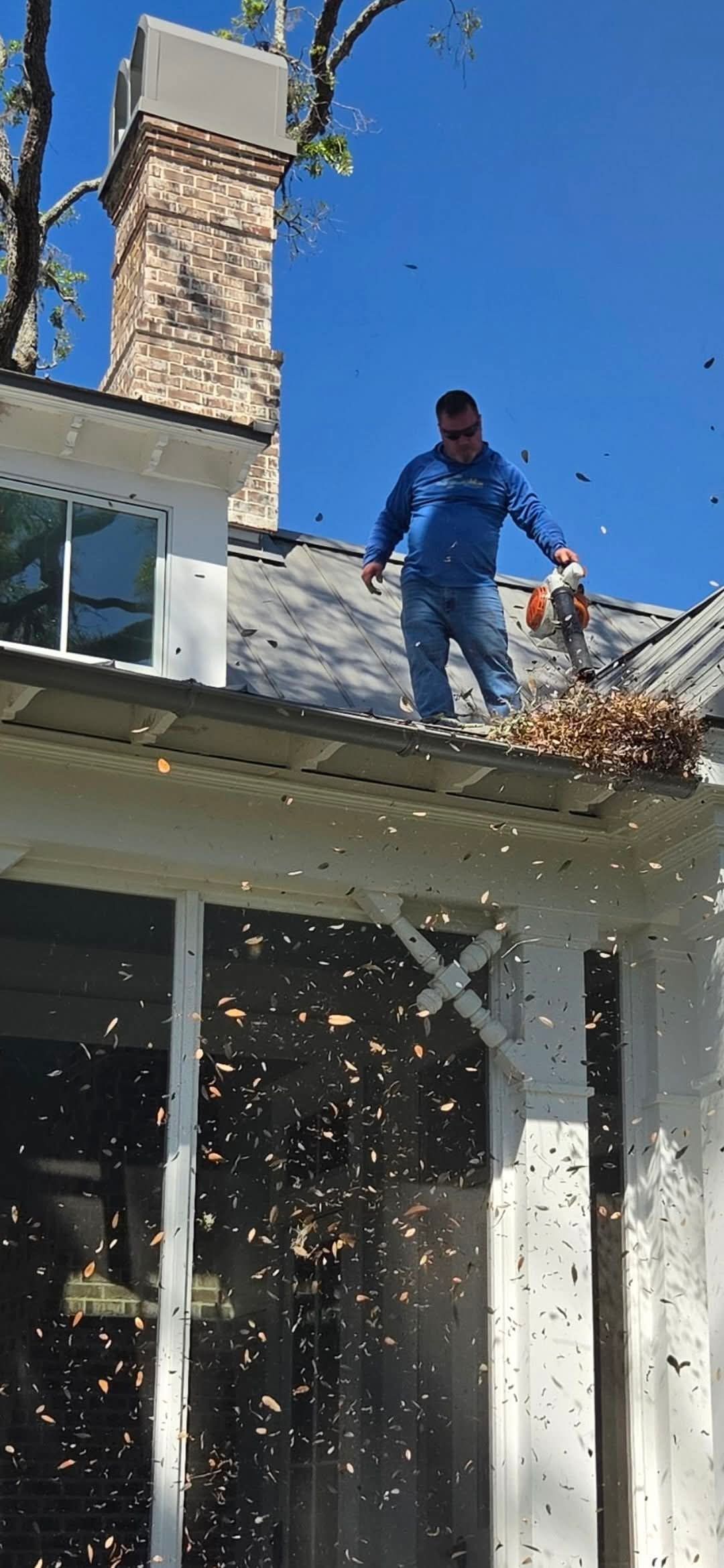 White gutter downspout under a house roof eave with perforated soffit and blue sky visible above tree tops