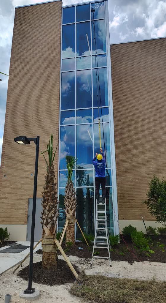 Worker on a ladder cleaning a glass tower window beside a brick building