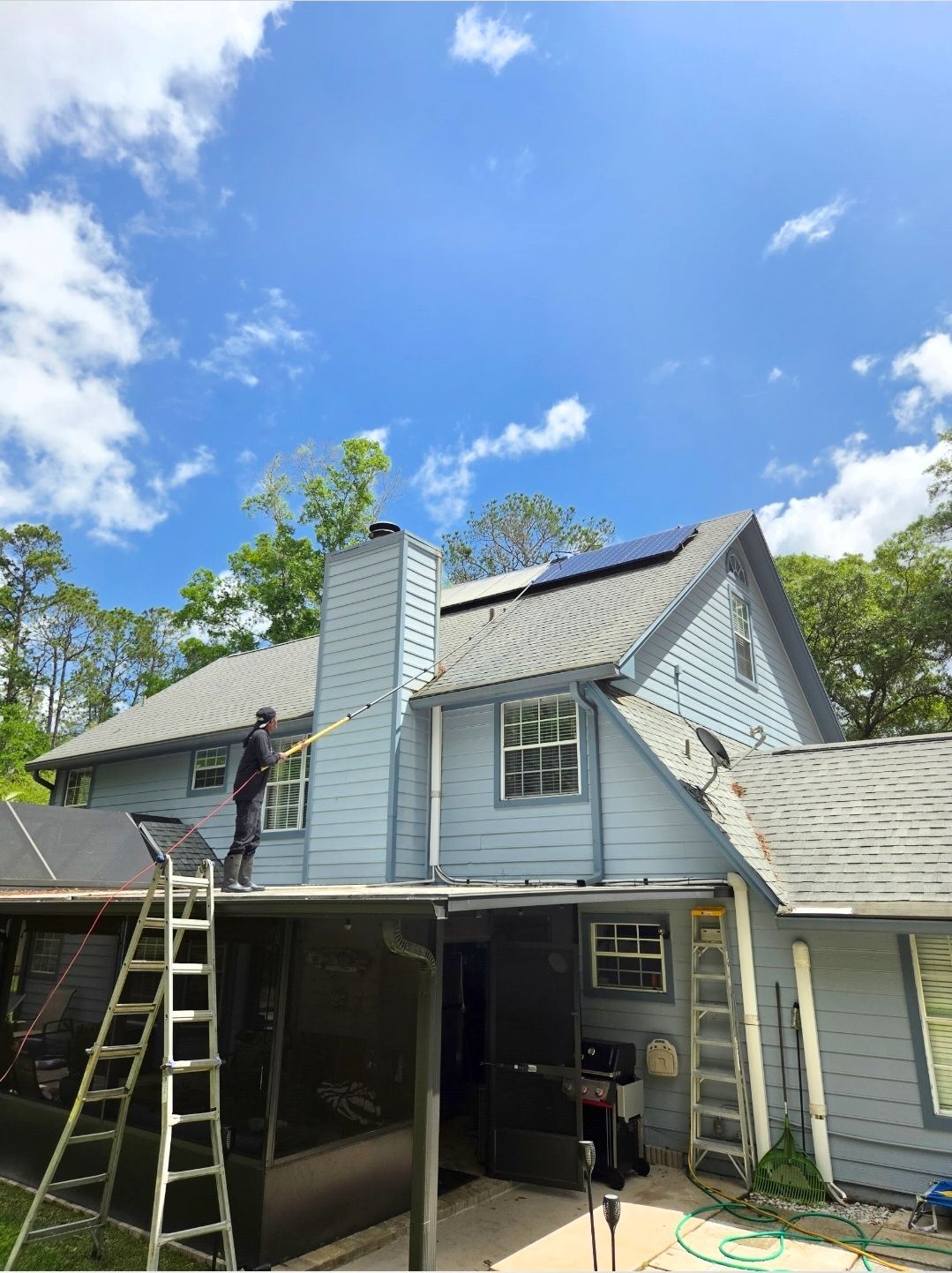 Two-story light-blue house with roof repair, ladder, and open garage under a blue sky