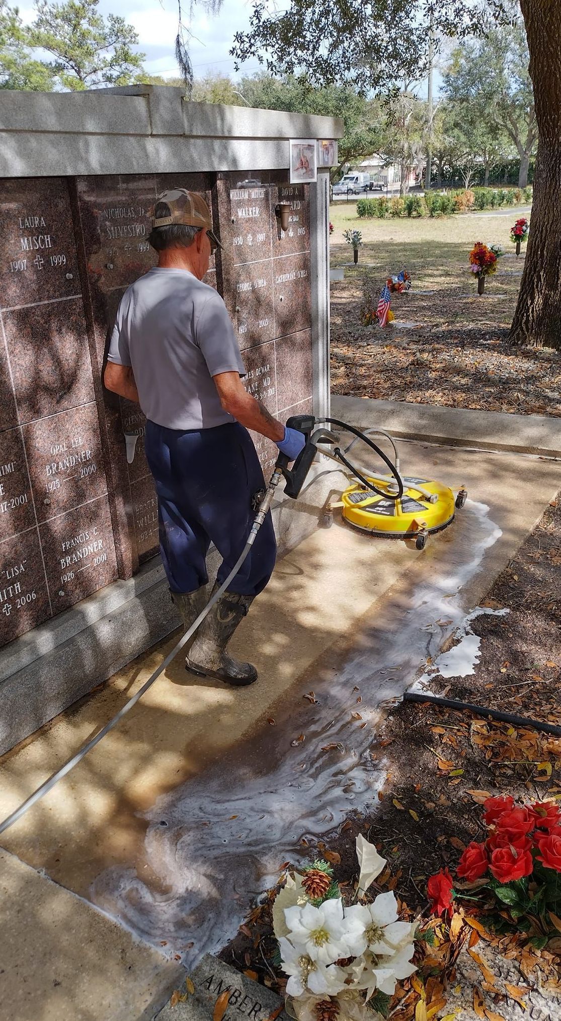 Person pressure-washing a stone walkway beside a building, with flowers in the foreground.