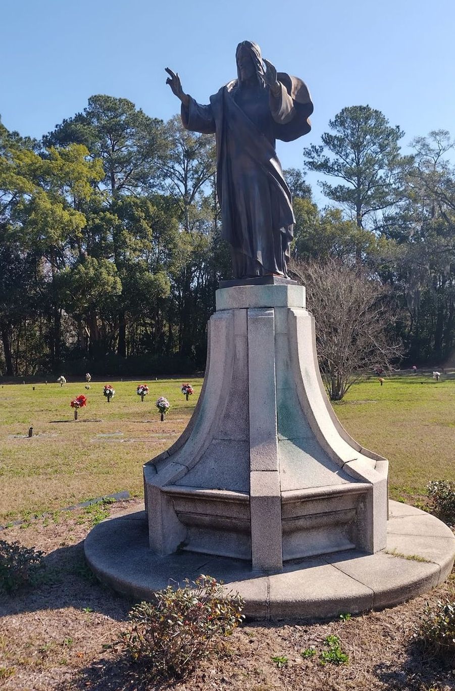 Statue of a robed figure on a pedestal in a grassy park, with trees in the background.