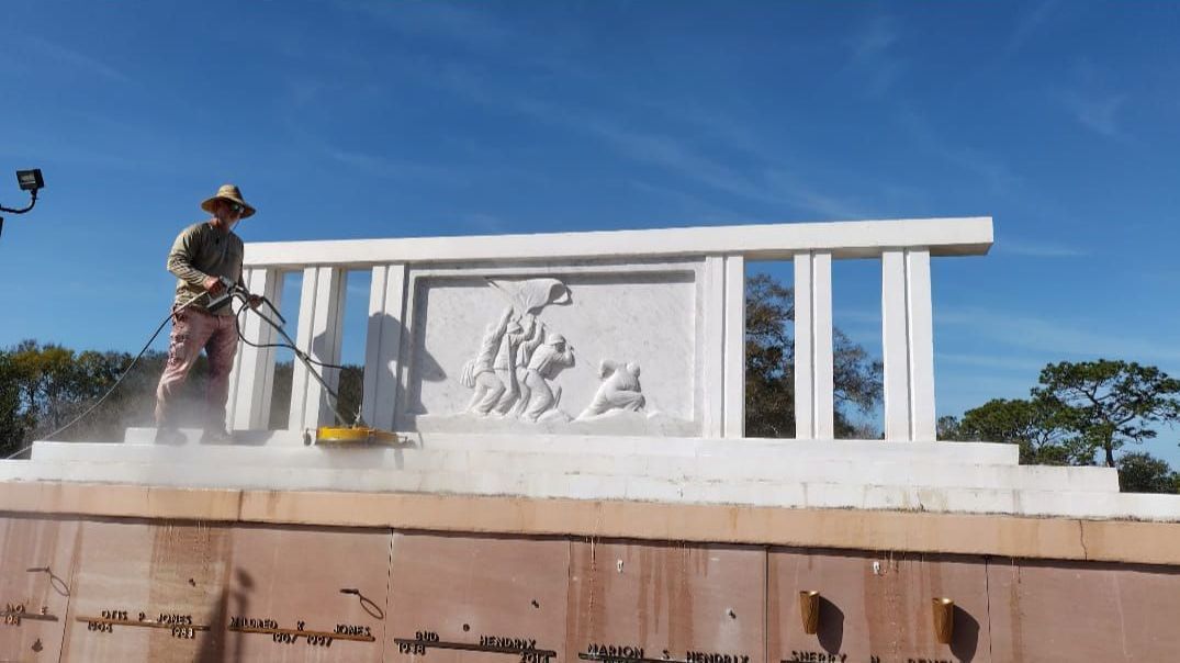Worker spray-cleaning a white memorial monument under a clear blue sky