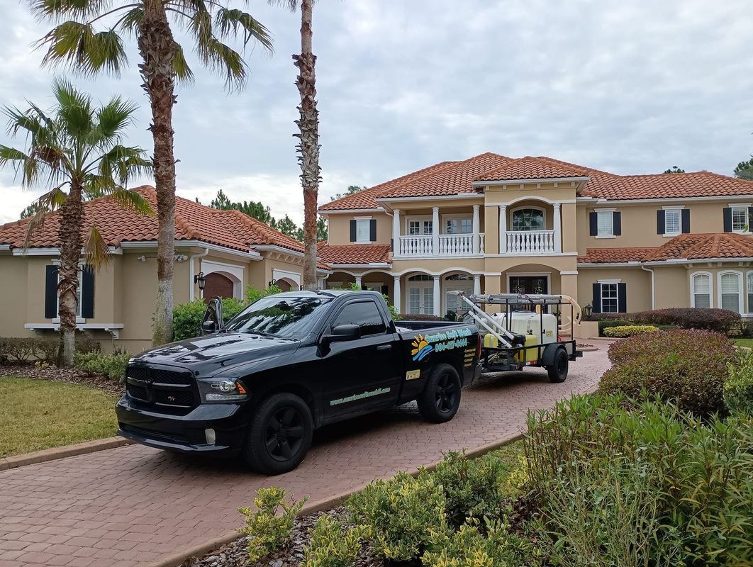 Black tow truck hauling a trailer in front of a large Mediterranean-style house with palm trees.