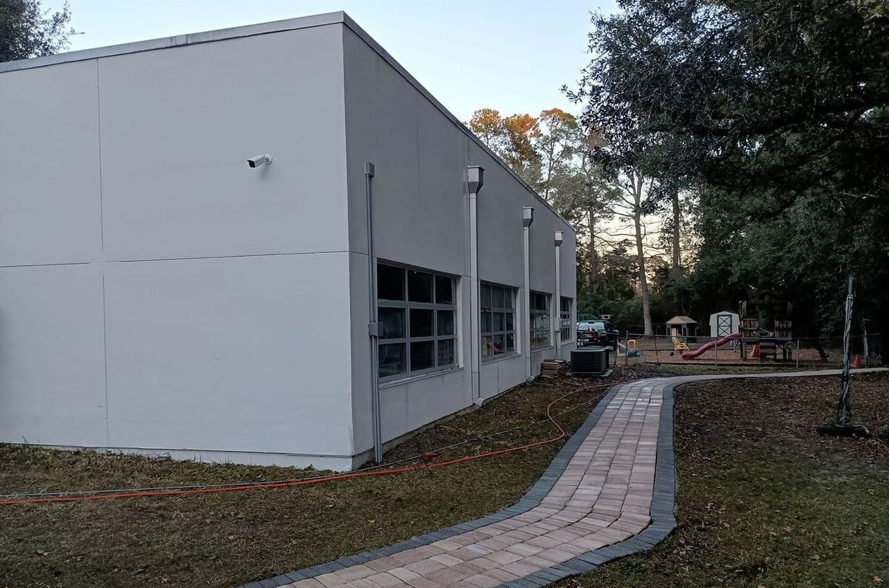 White building exterior with windows, a paved walkway, and trees in a grassy yard.