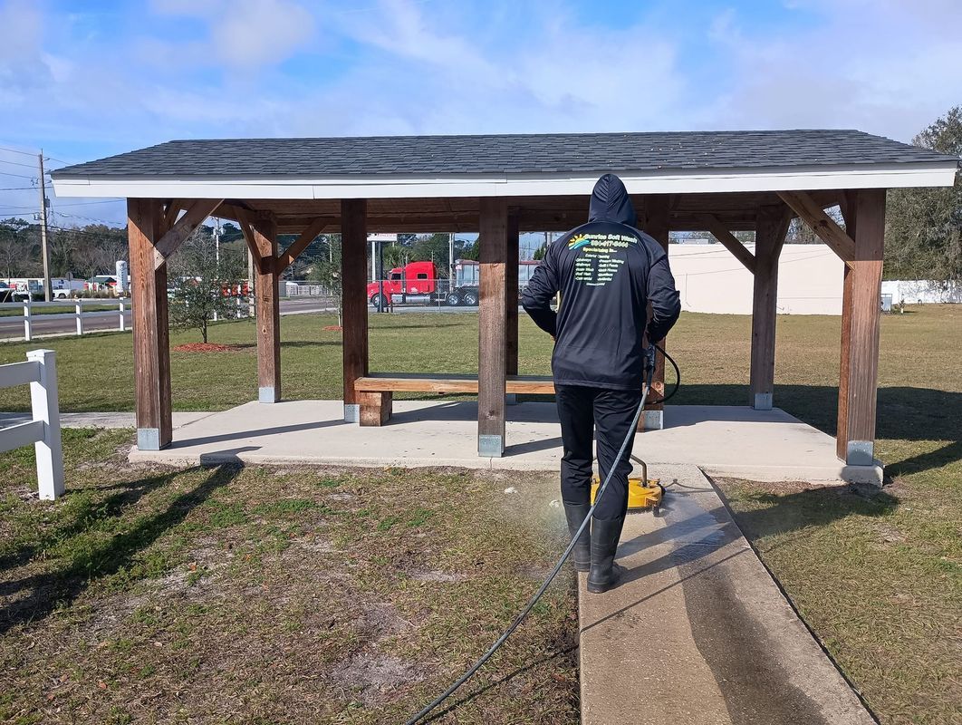 Person in a black jacket walking under a small wooden pavilion on a sunny day.