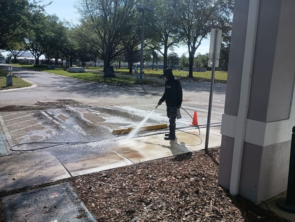 Person spraying water on a wet sidewalk beside a building, with an orange cone nearby.
