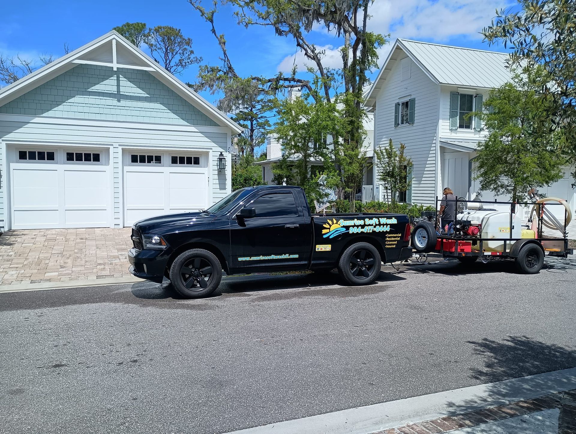 Black pickup truck towing a red utility trailer parked in front of a white garage on a sunny street