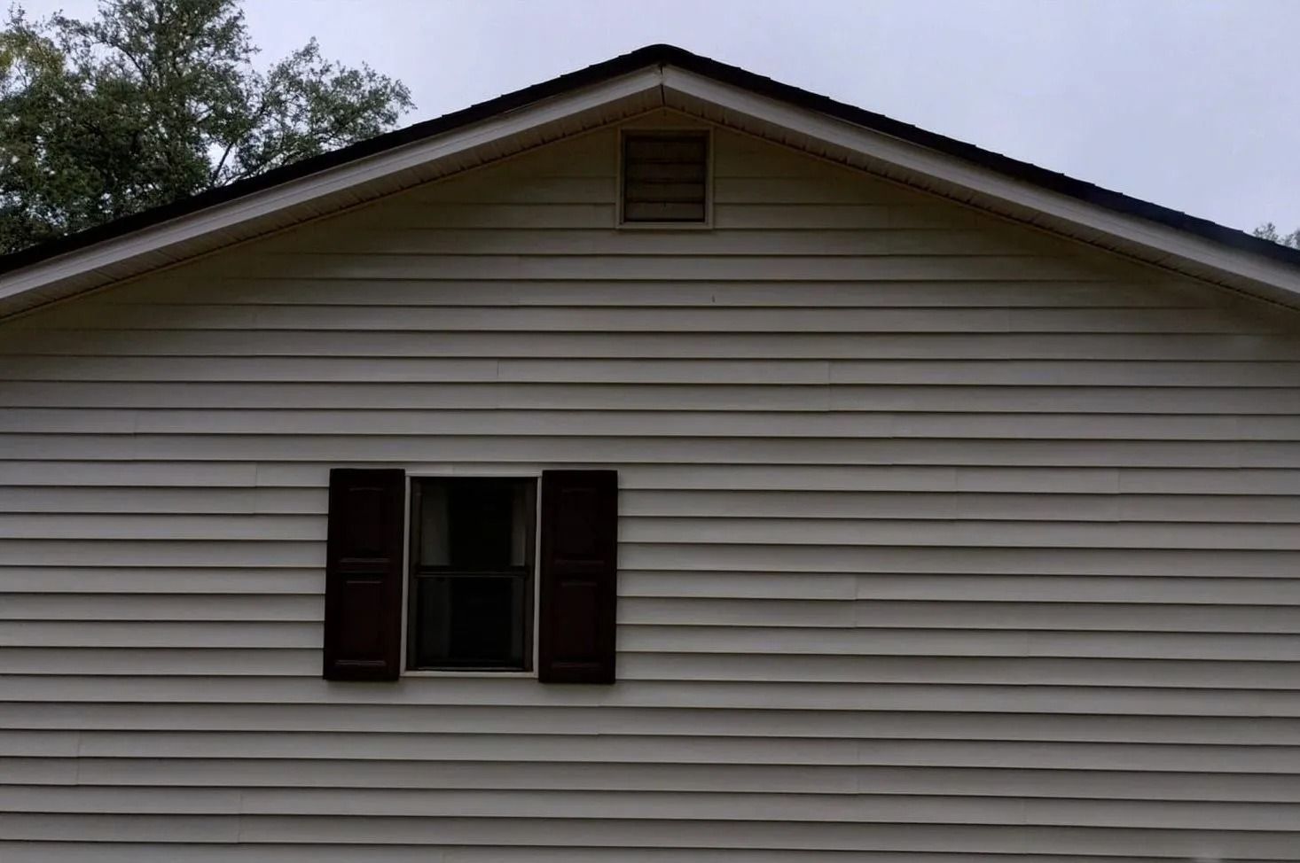 Front of a white house with horizontal siding and a centered gable vent, with dark shutters around a window.