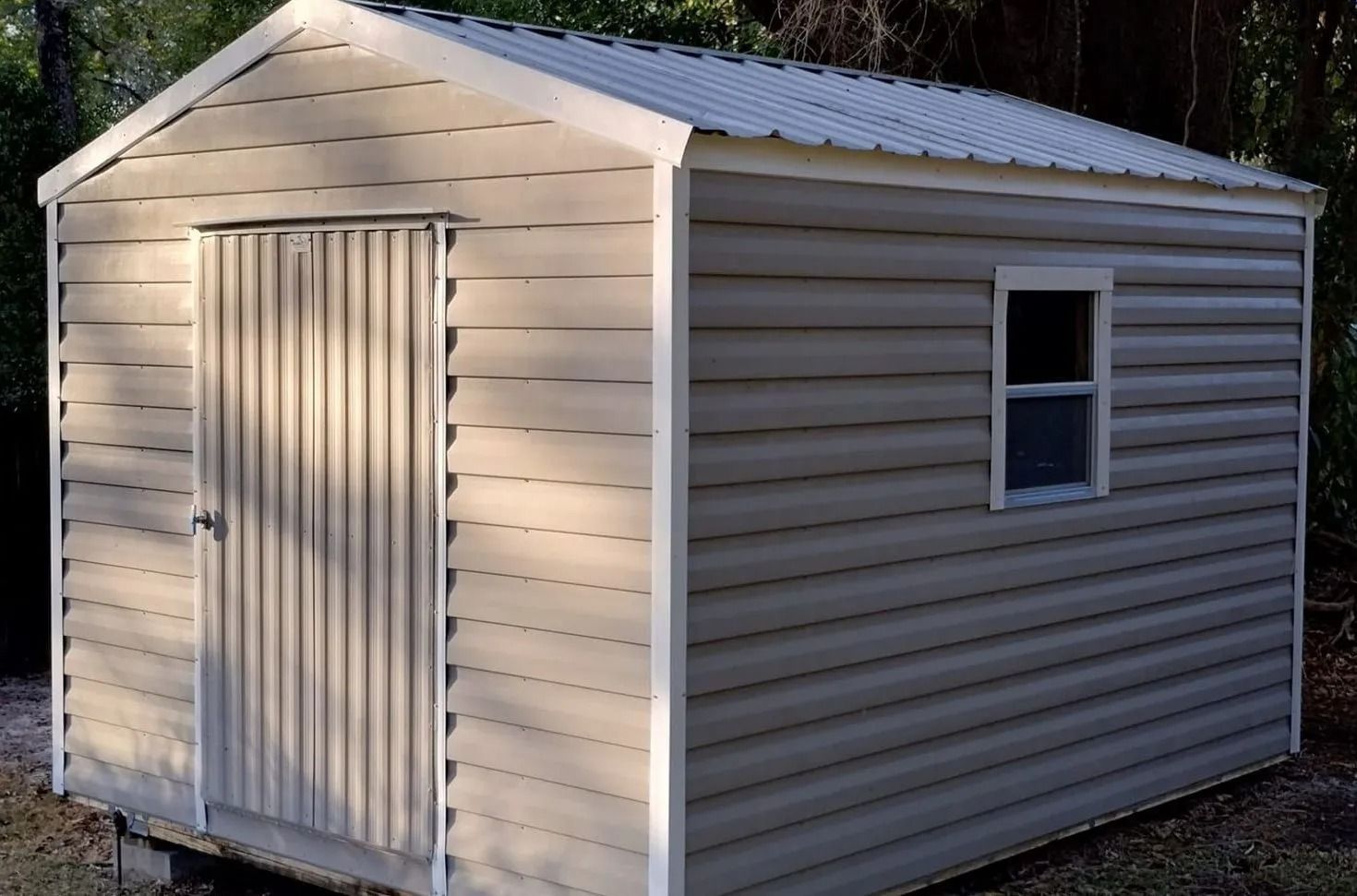 Small beige storage shed with a white door and one window, standing among trees outdoors