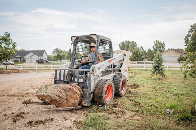 Worker operates a skid steer moving a large rock on a dirt construction site.