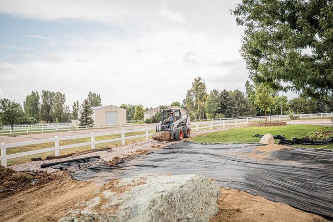 Worker operating a skid steer on a dirt driveway beside a white fence and trees