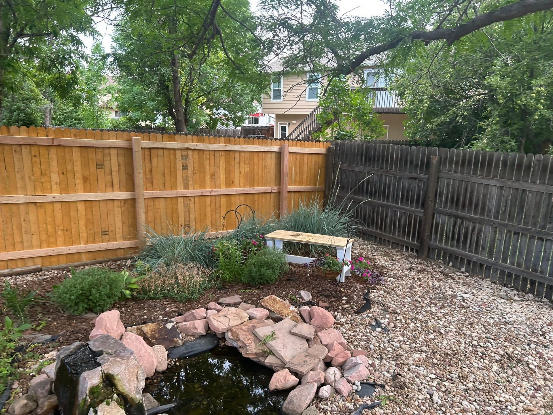 Backyard garden with pond, rock border, flower bed, and wooden fences under leafy trees