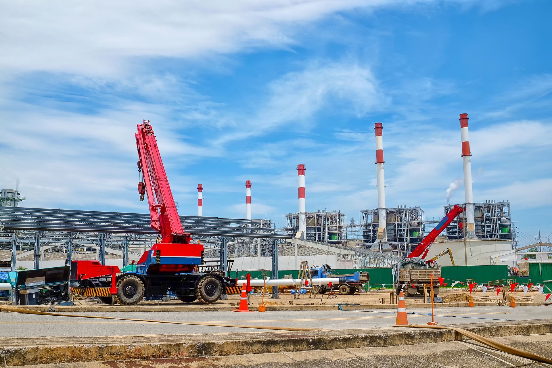 Construction site with red cranes, industrial structures, and smokestacks under a blue sky