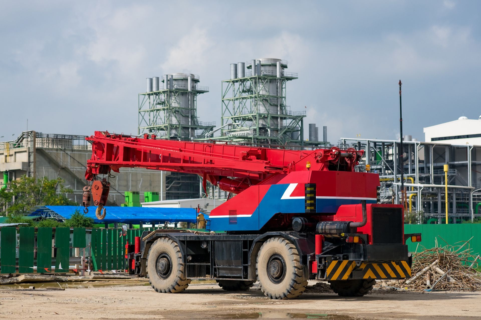 Red telescopic crane truck at a construction site with industrial tanks in the background