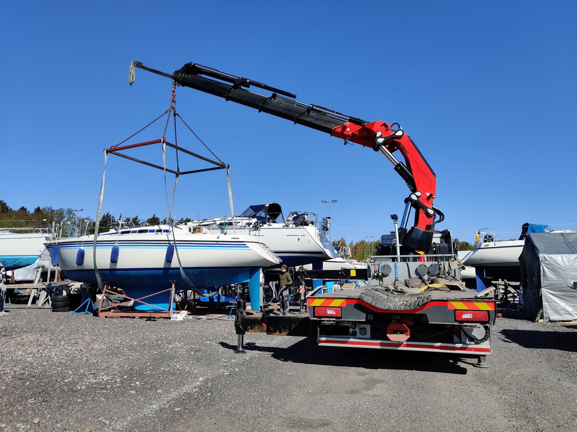 Crane lifting a white sailboat on a trailer in a boatyard under a blue sky