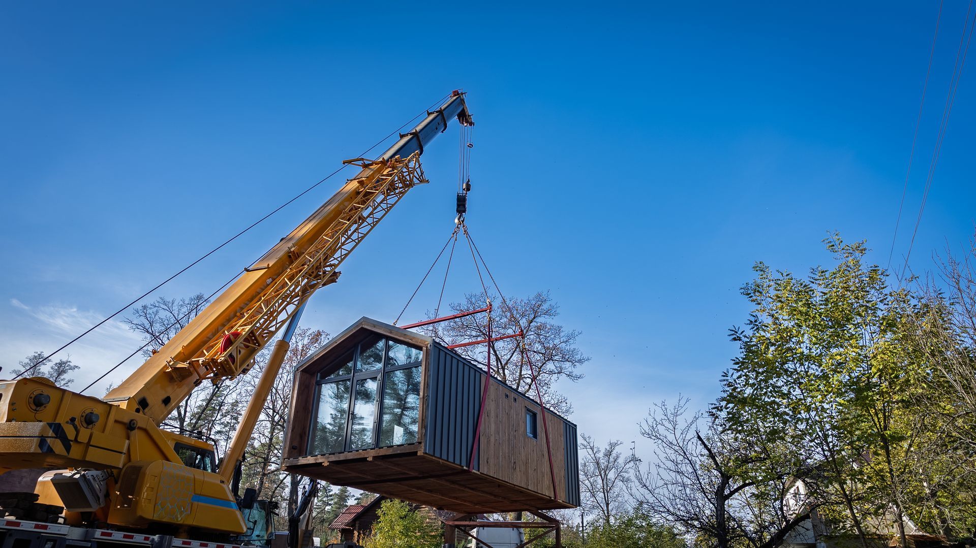 Yellow crane lifting a small modular house against a blue sky and trees