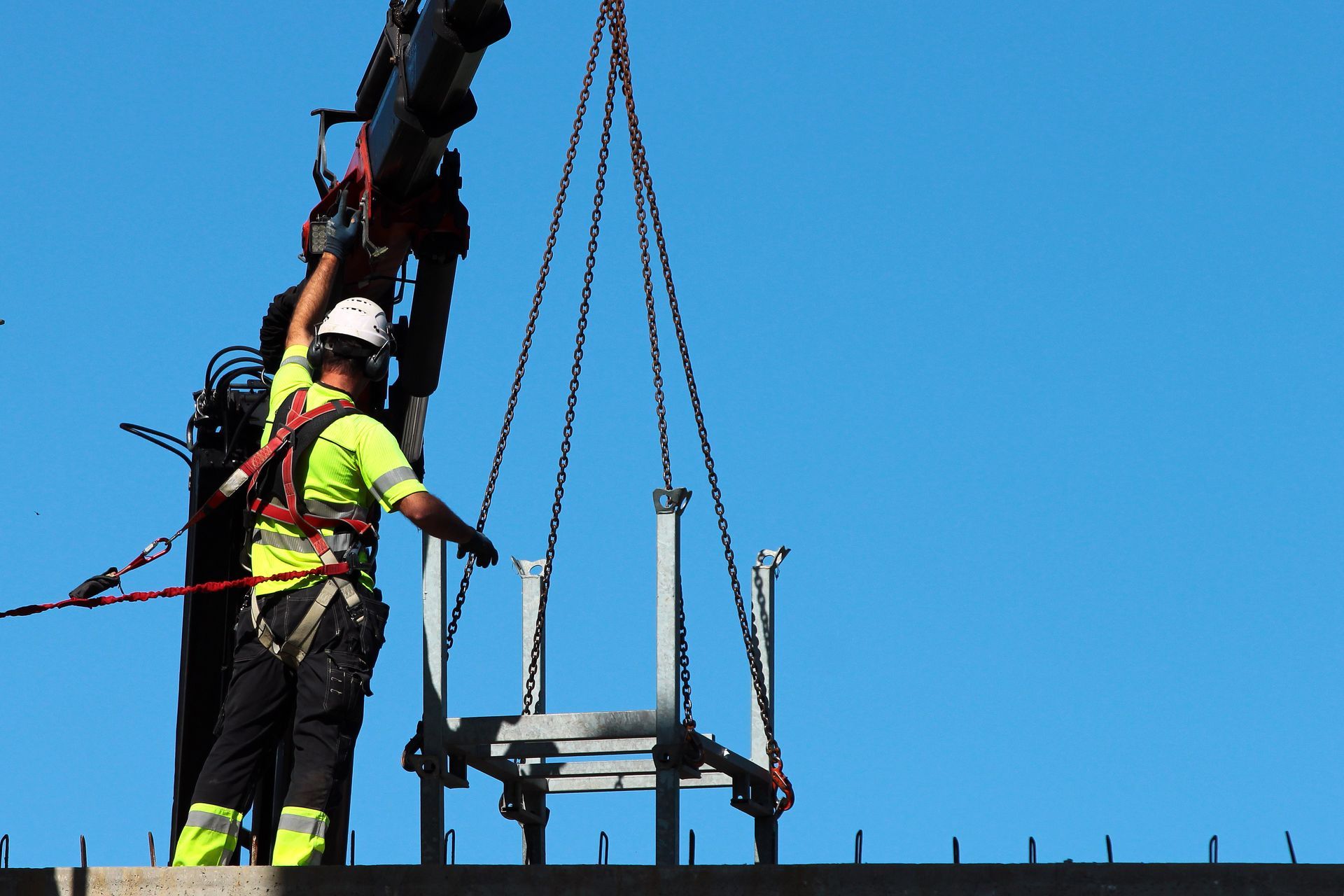 Worker in a neon vest and hard hat guiding a suspended platform crane lift against a blue sky