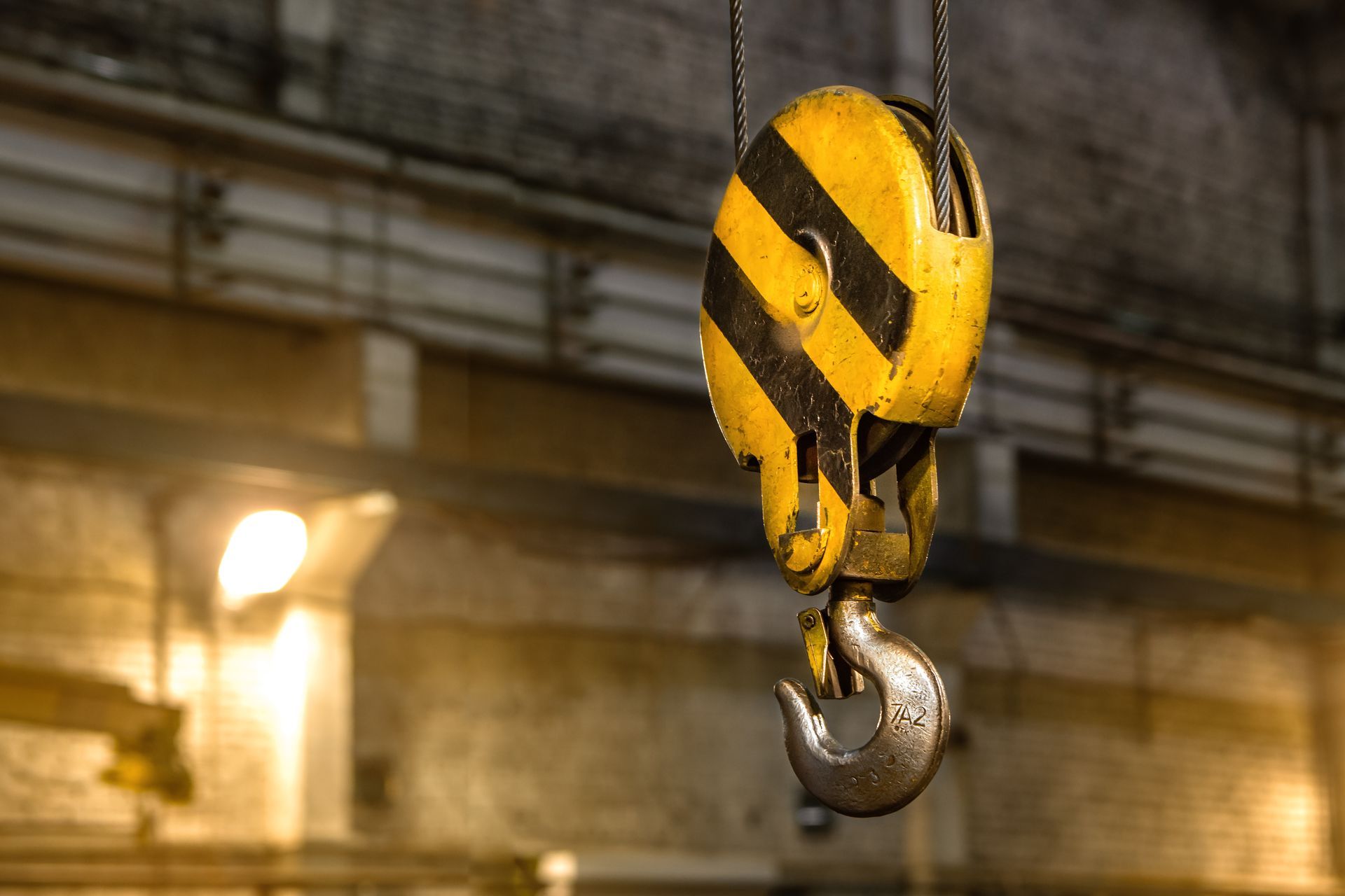 Yellow-and-black industrial crane hook hanging in a warehouse with blurred background lighting