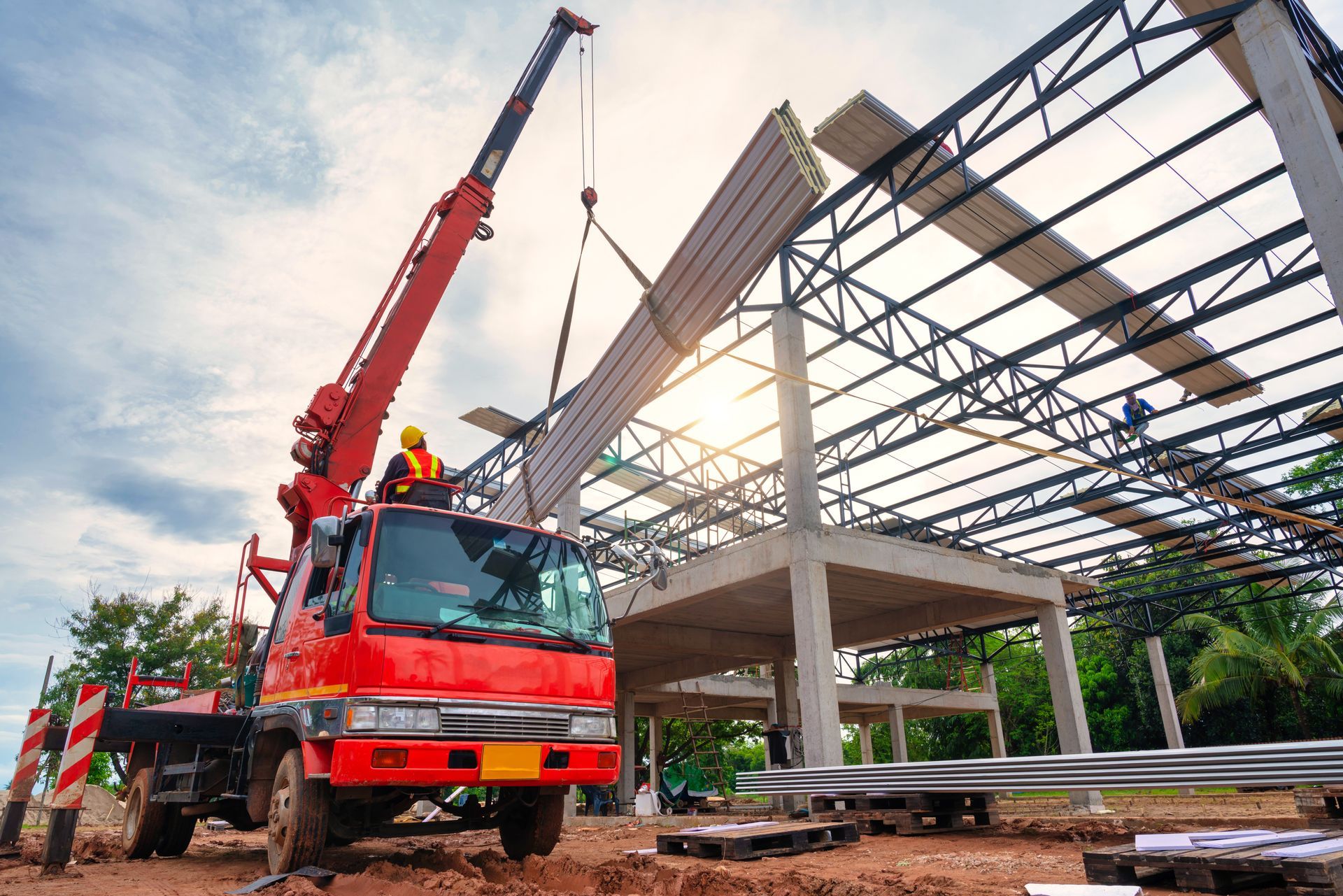 Red crane lifting steel beams on a construction site with workers and a partially built structure