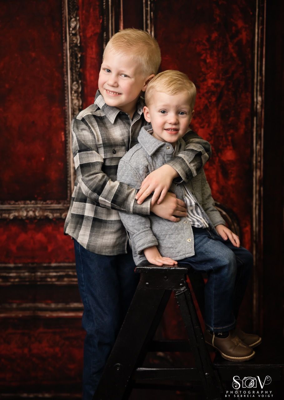 Two young boys smiling, embracing in front of a red backdrop; one on a small step stool.