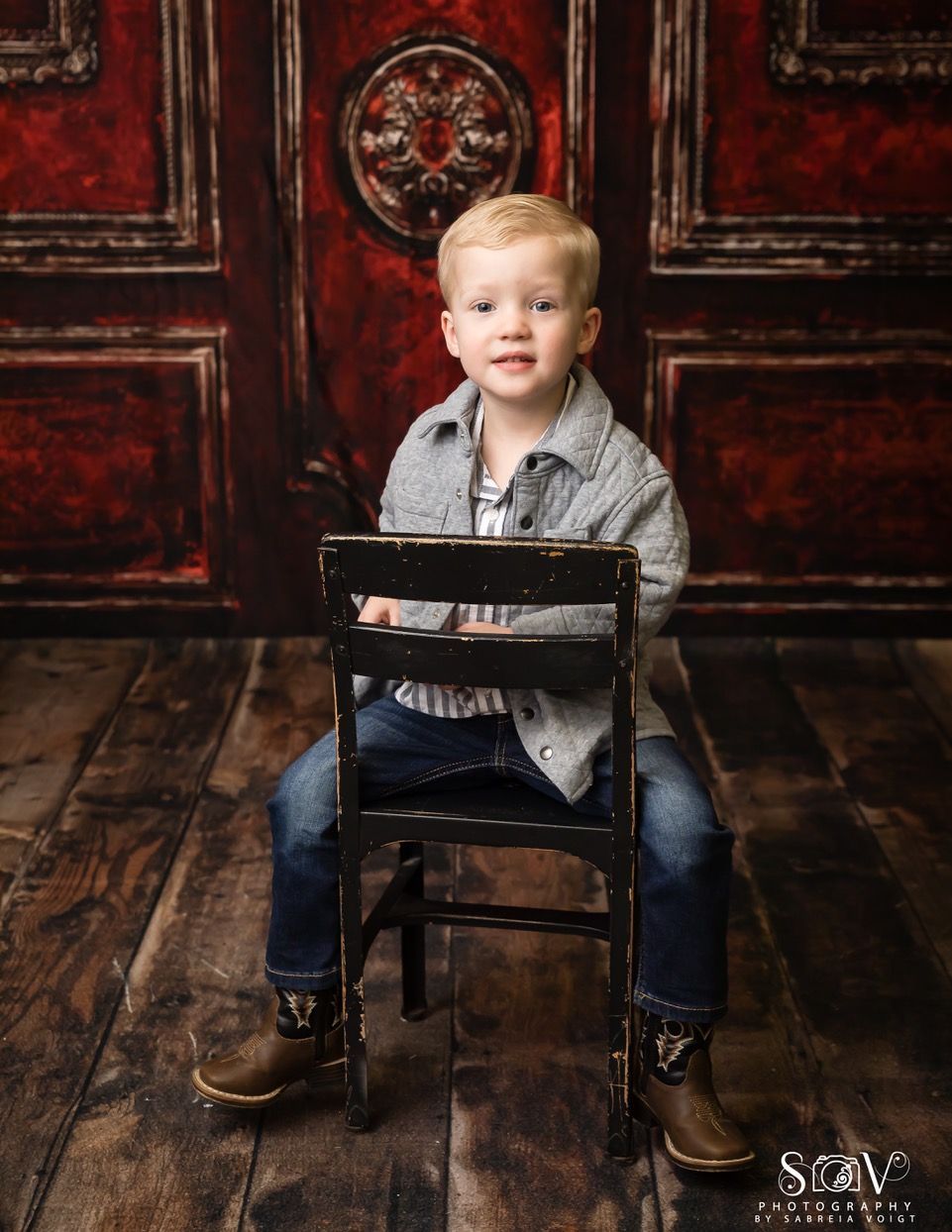 Boy in denim jacket and jeans sits on a chair, western boots on wooden floor, red background.