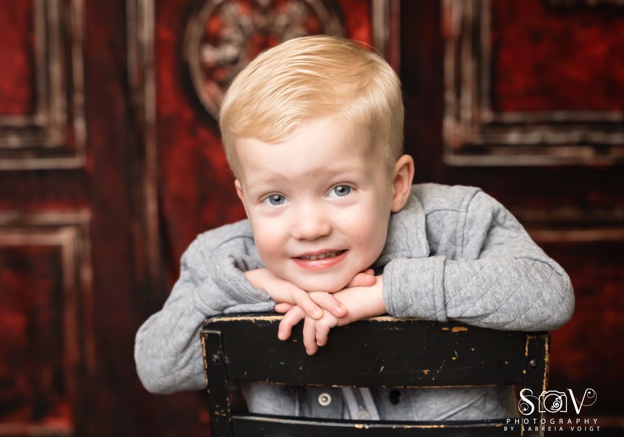 Blond-haired child with blue eyes, smiling, resting arms on a chair, red backdrop.