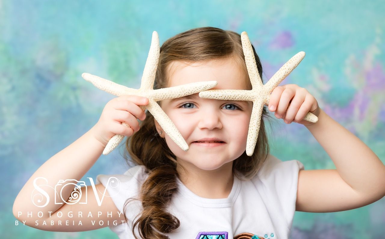 Young girl smiling, holding two starfish near her face against a blue and purple background.