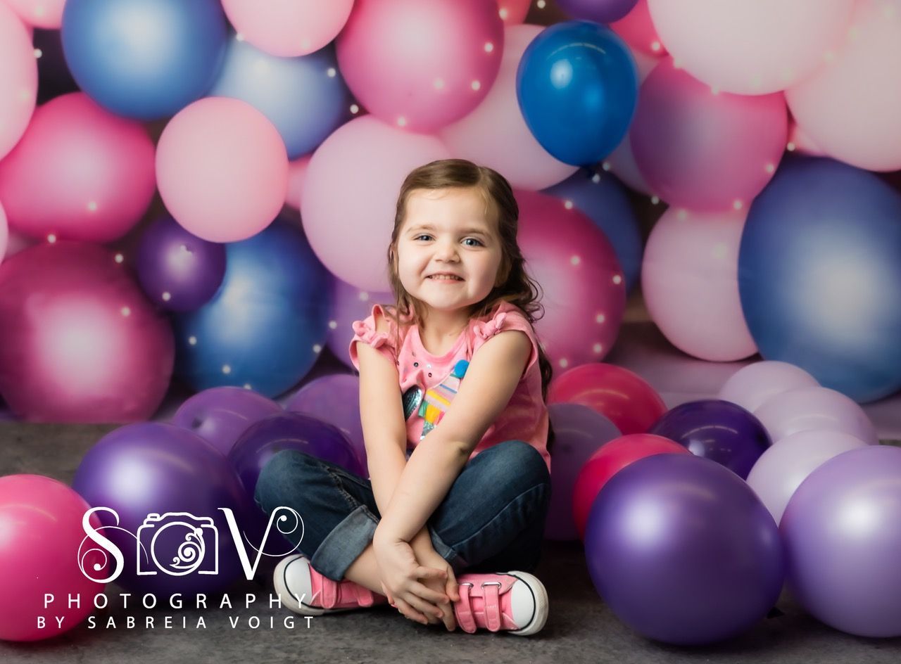 Smiling child sits surrounded by pink, purple, and blue balloons.