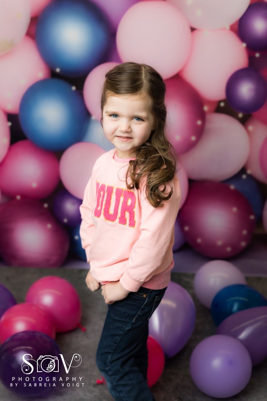Girl in pink sweatshirt, standing in front of colorful balloons.