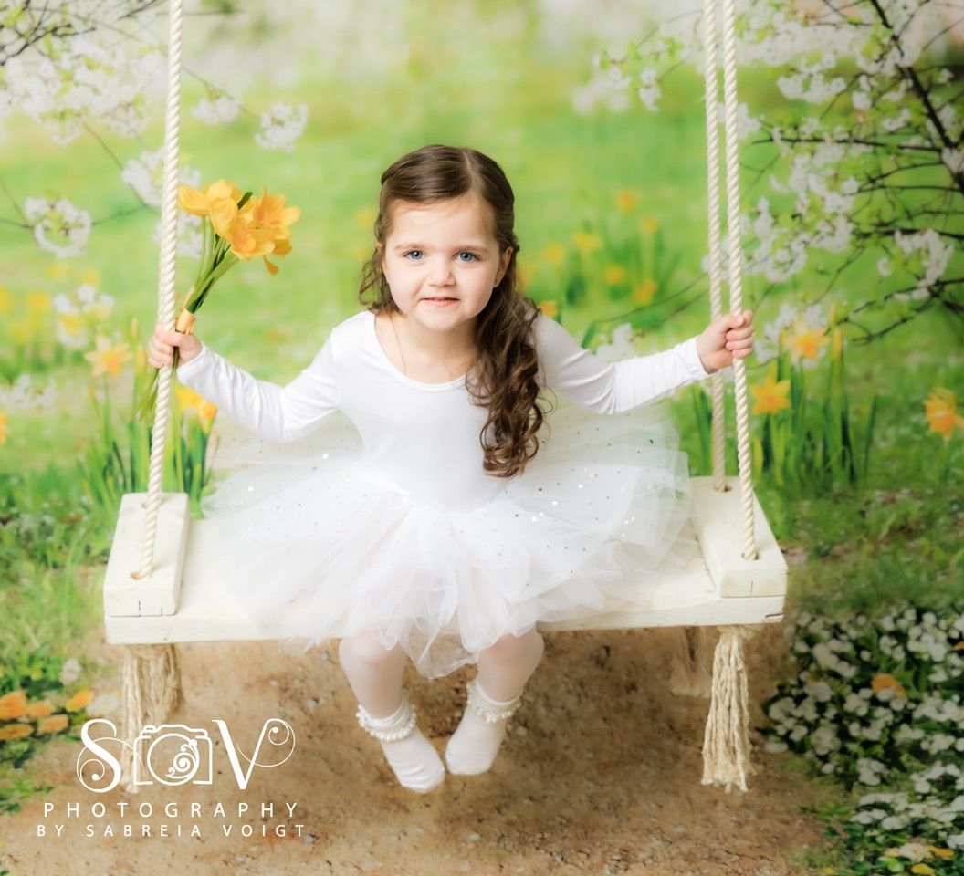 Girl in white tutu on a swing, holding yellow flowers, in a spring garden.