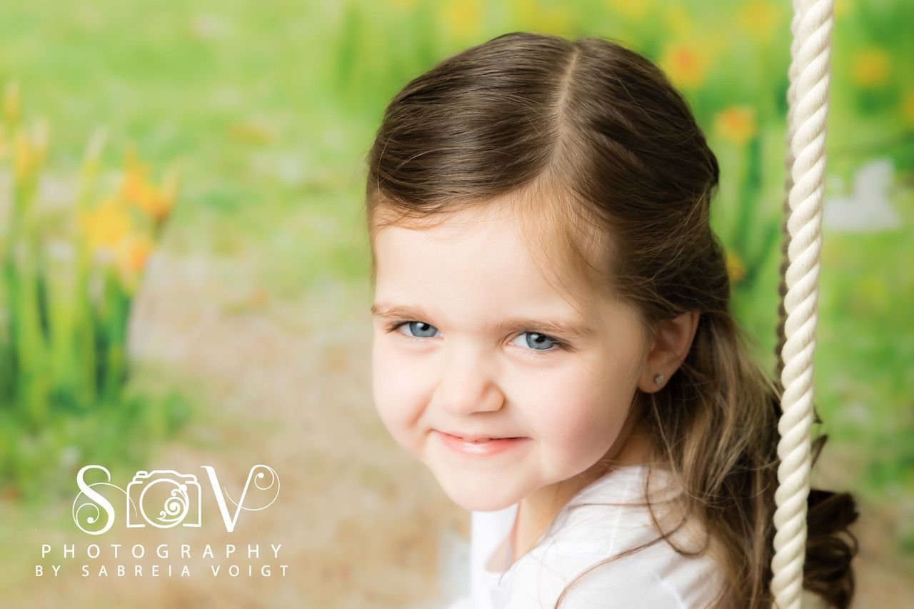 Young girl with blue eyes smiles, seated on a swing, in front of a green floral backdrop.
