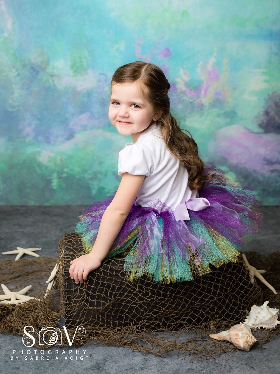 Girl in mermaid-themed tutu, smiles, poses on fishing net with seashell and starfish, backdrop with sea colors.